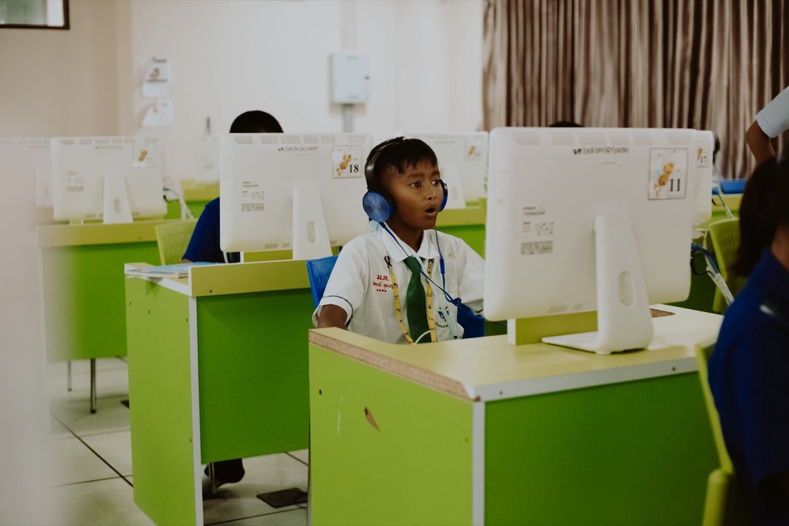 A boy in a white school shirt wears headphones while working at a computer in a classroom