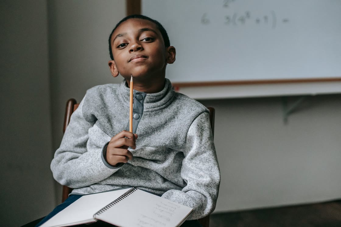 A schoolboy in a grey jumper sits in front of a whiteboard, holding a pencil to his chin and thinking, with an open notebook on his lap