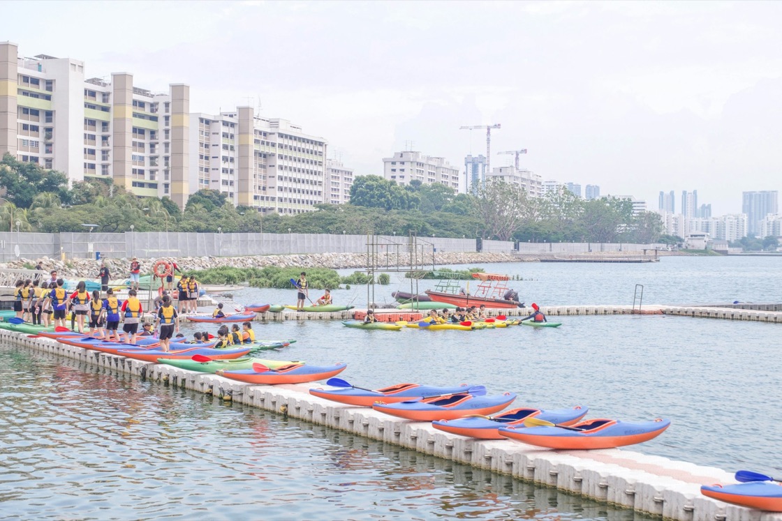 Young people in life jackets prepare to launch kayaks from a floating dock, with flats and the city skyline behind them