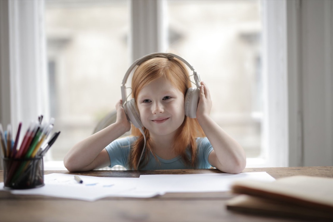 A smiling girl sits at a desk with her hands on a pair of large headphones, with an open notebook in front of her