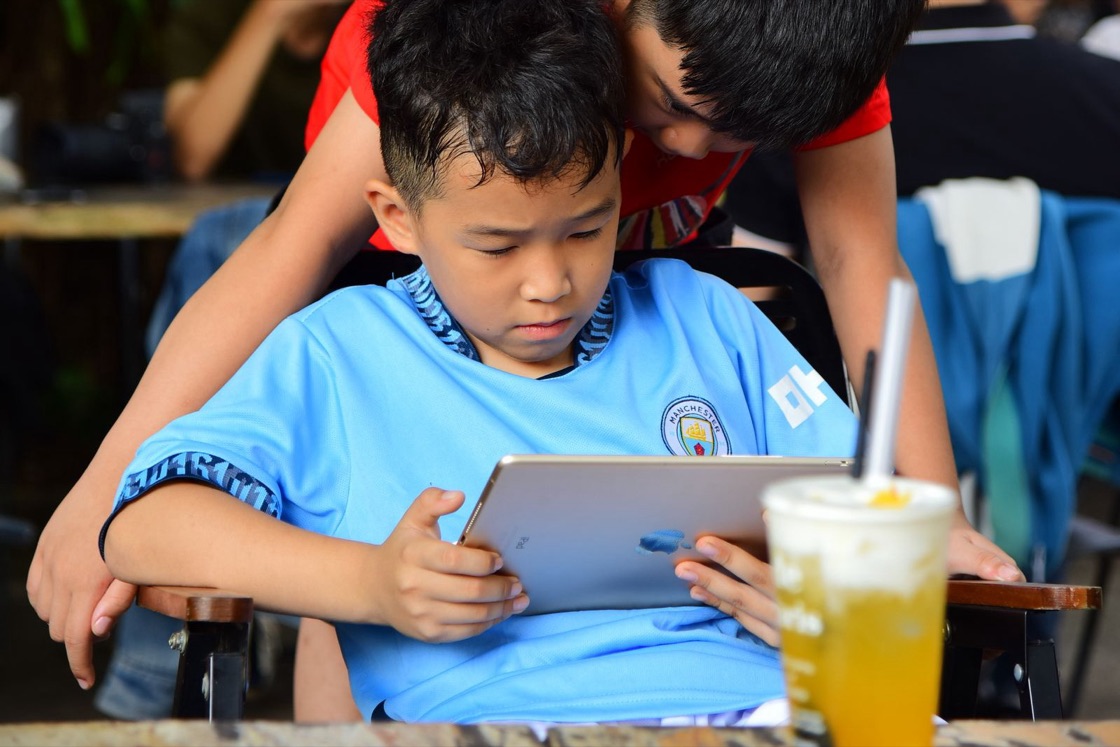 A boy looks closely at a tablet at a café table while an older boy leans over his shoulder to watch