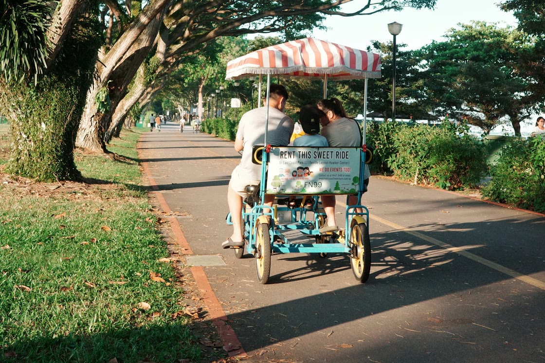 A family rides a four-wheeled surrey bike with a striped canopy along a shady path lined with rain trees at East Coast Park