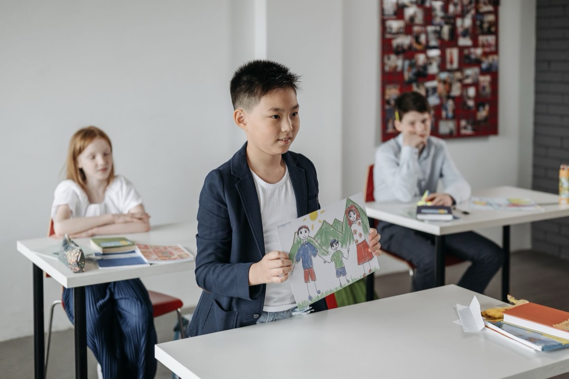 A boy stands at the front of a classroom and shows his classmates a drawing of his family