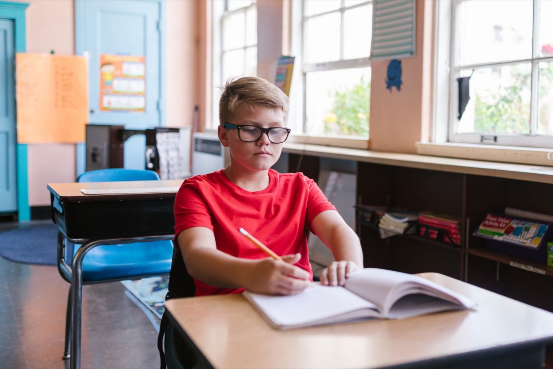 A boy in glasses and a red T-shirt sits at a classroom desk, writing in an open book with a pencil