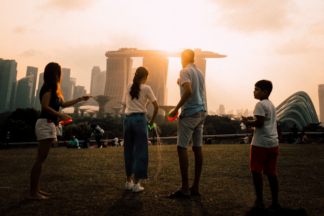 A family flies kites on a grassy field at sunset, with Marina Bay Sands and the Gardens by the Bay domes behind them