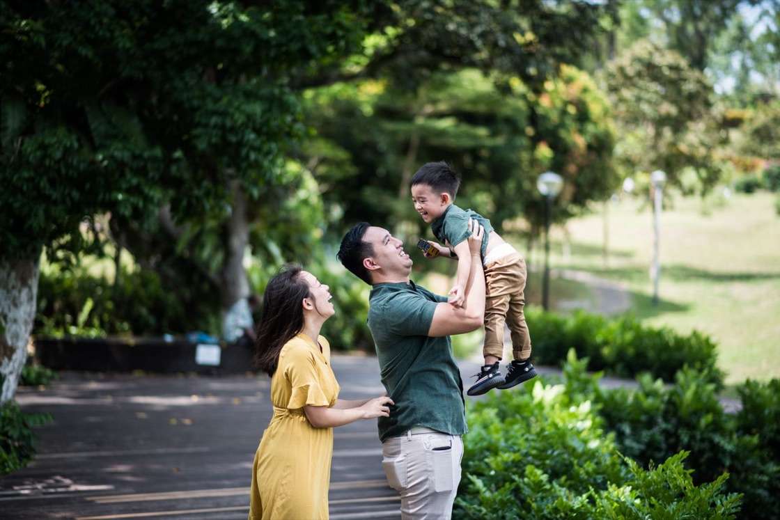 A father lifts his laughing young son into the air while the mother smiles beside them in a green park