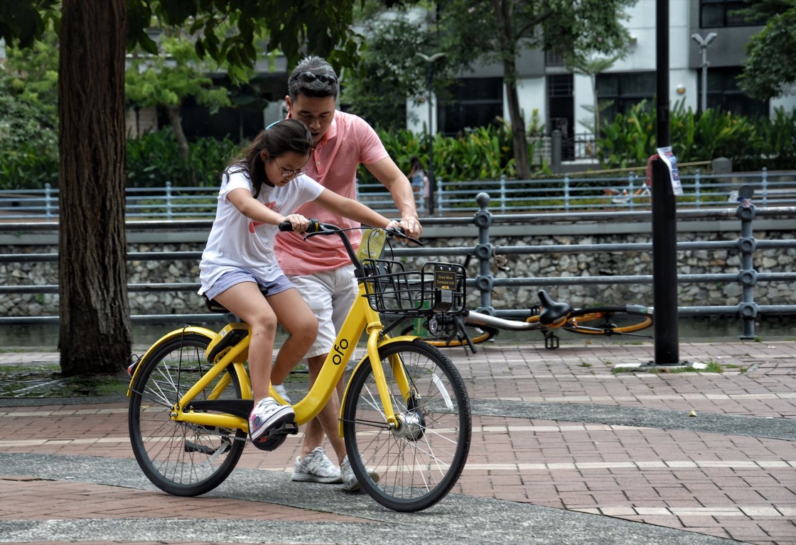 A father steadies a yellow shared bicycle while his daughter pedals along a park path, with another bicycle lying on the ground behind them