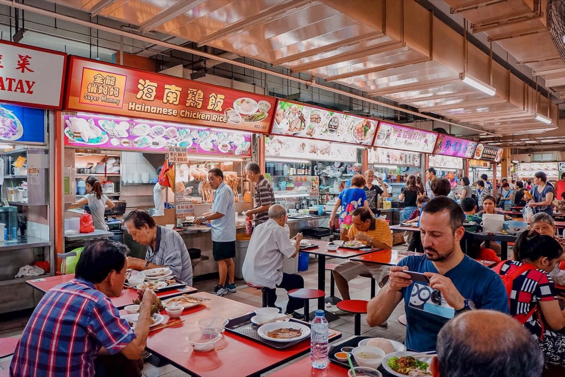 Diners of different ages eat at crowded tables in a Singapore hawker centre, with a chicken rice stall behind them