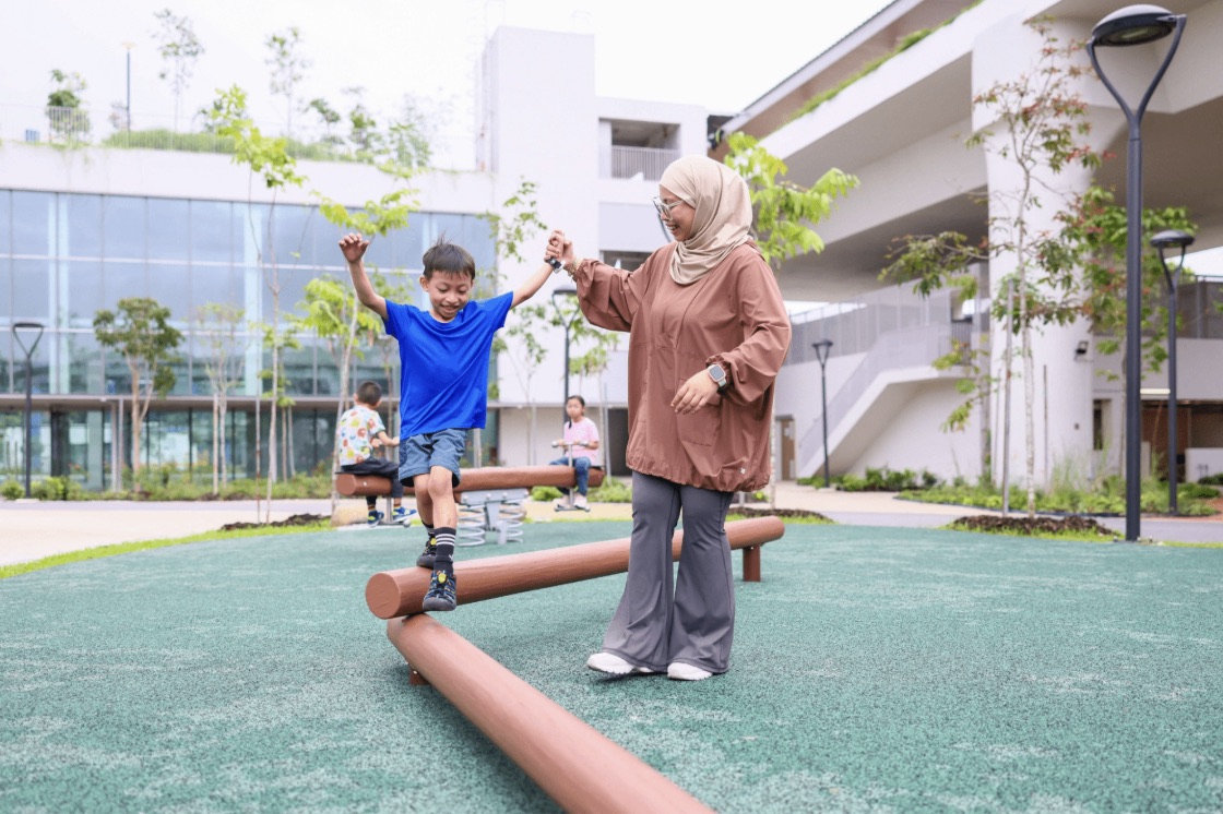 A boy balances on a low wooden beam at a neighbourhood playground while his mother holds his hand, with other children playing behind them