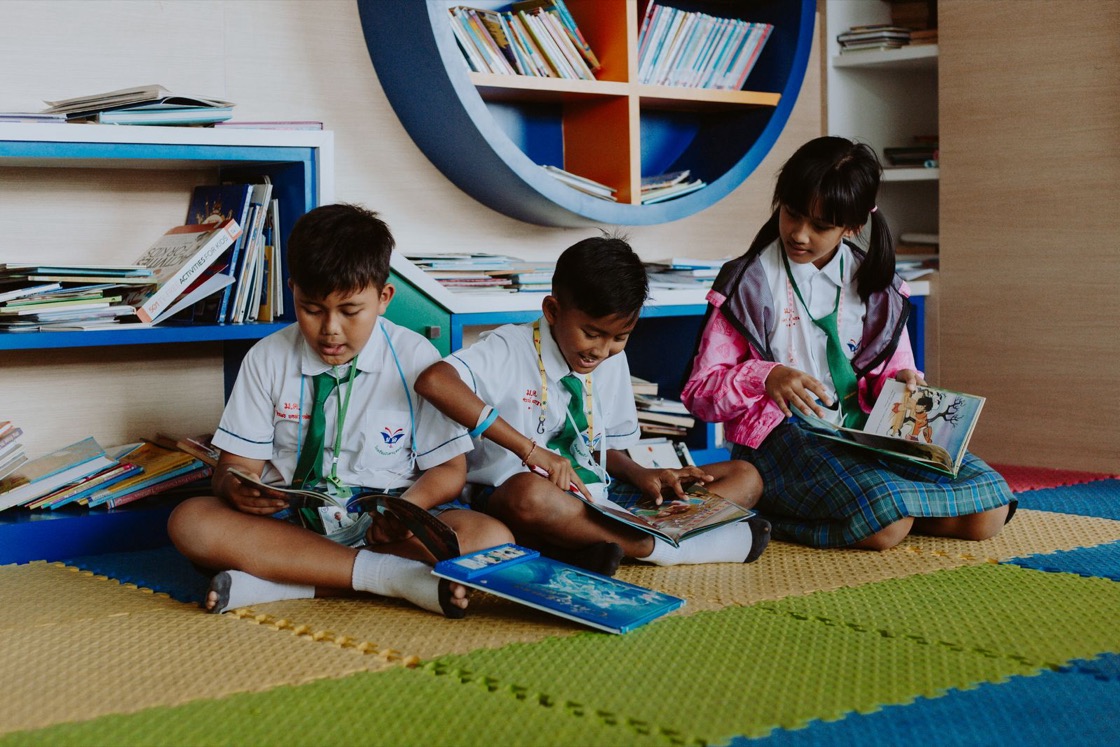 Three primary school children in uniform sit on a library floor, each reading a book