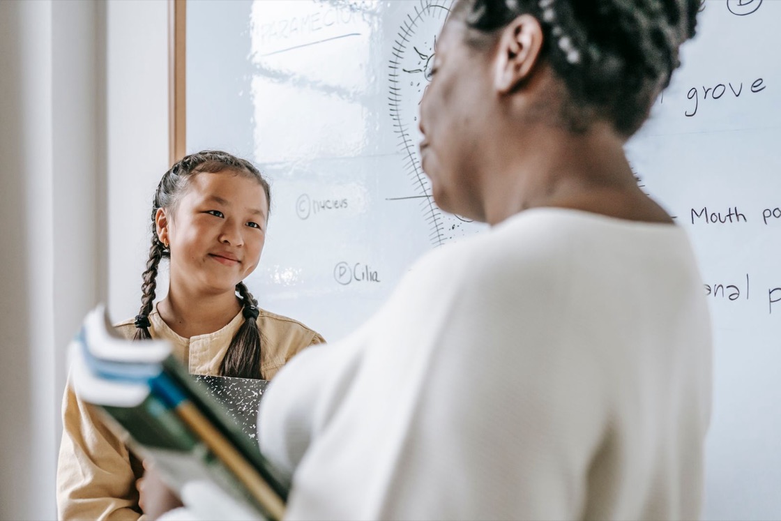 A smiling girl holding books talks with her teacher in a classroom