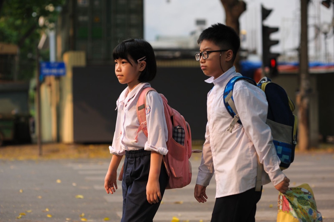 A girl and a boy in white school shirts, carrying backpacks, walk side by side along a city street on their way to school