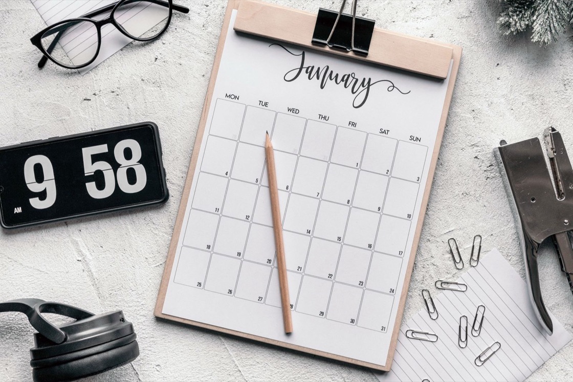 A blank monthly calendar on a clipboard, lying on a desk beside a pencil, glasses and paper clips