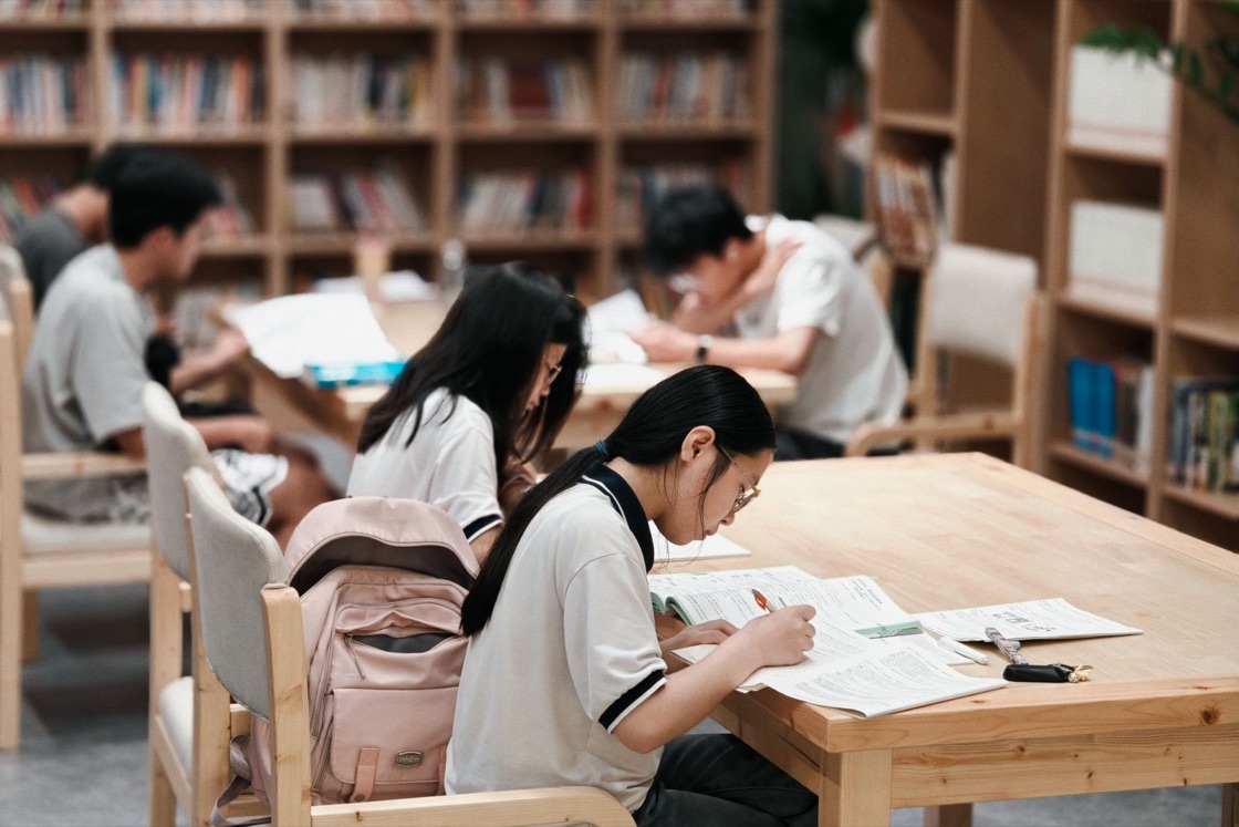 Students sit at wooden tables in a bright library, reading and writing notes from open books