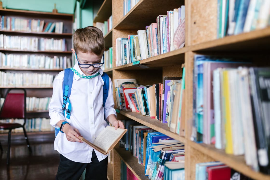 A boy in a white school uniform and glasses reads an open book while standing between tall library bookshelves