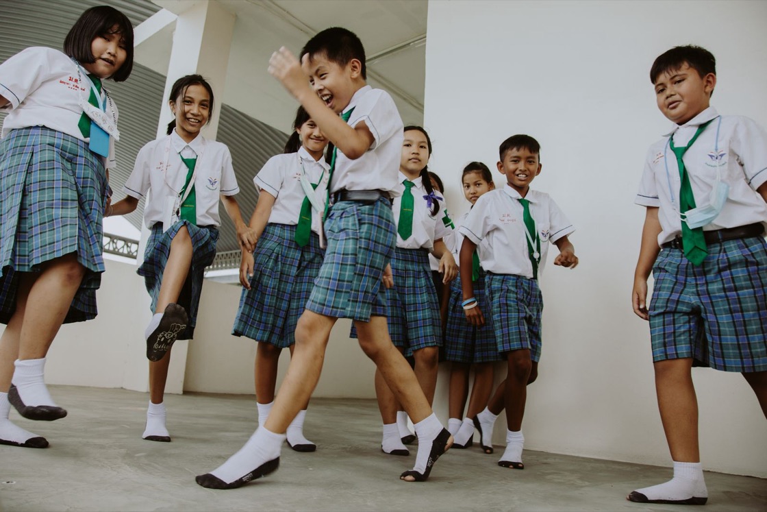 Children in checked school uniforms laugh and dance along a school corridor