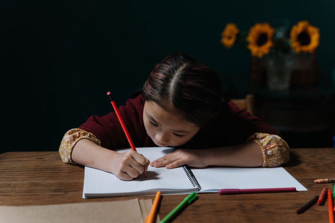 A girl leans over an open notebook at a wooden table, writing carefully with a pencil